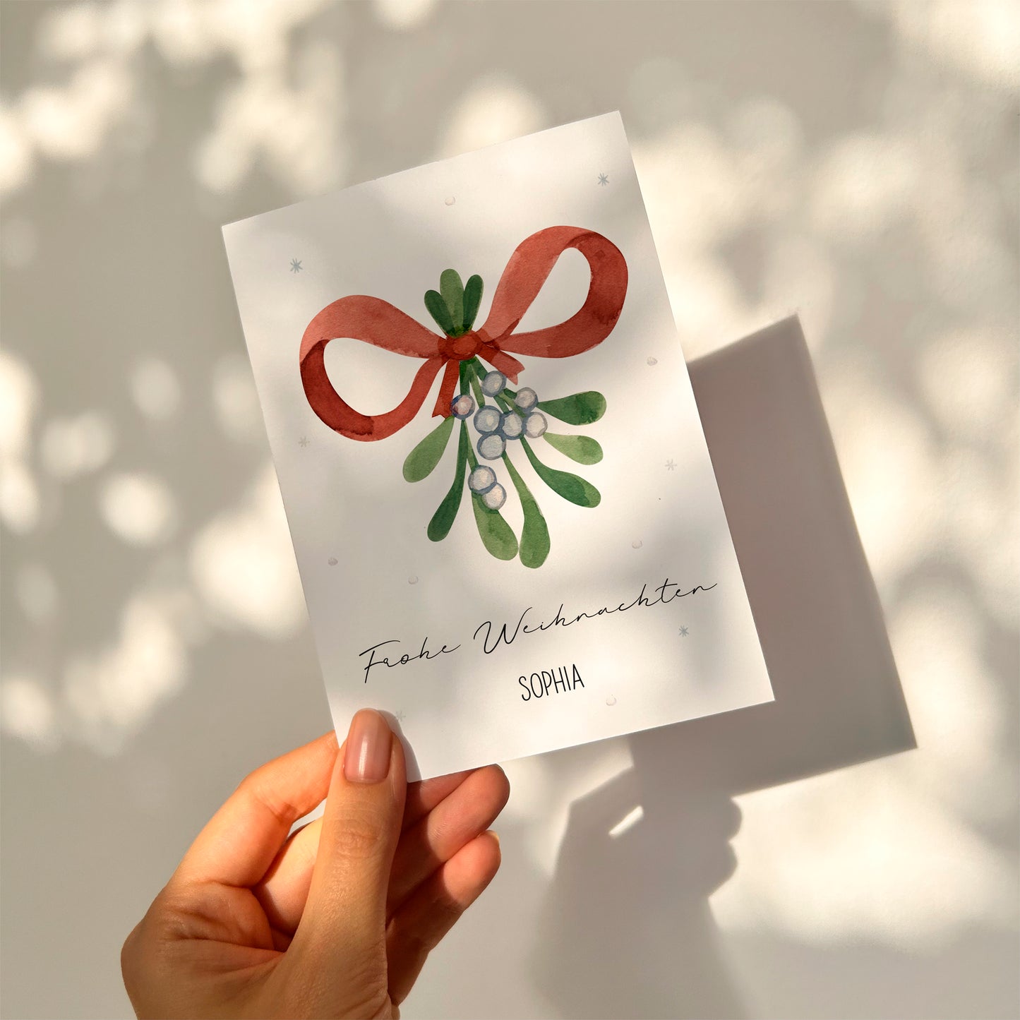 Hand hält Personalisierte Weihnachtskarte Mistelzweig A6 mit Aquarell-Motiv von Mistelzweigen, rotem Band und Text „Frohe Weihnachten“ sowie Name „Sophia“.