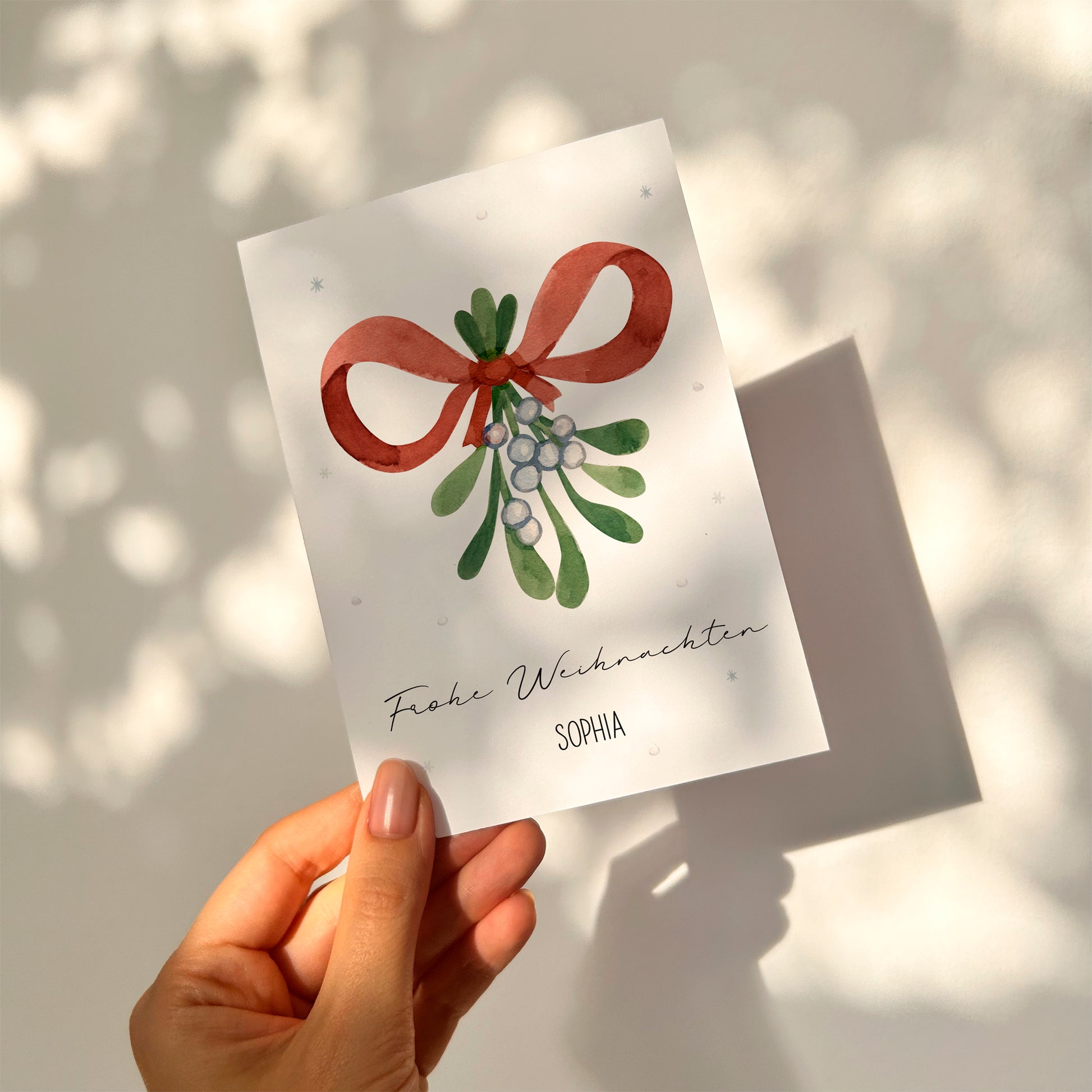 Hand hält Personalisierte Weihnachtskarte Mistelzweig A6 mit Aquarell-Motiv von Mistelzweigen, rotem Band und Text „Frohe Weihnachten“ sowie Name „Sophia“.