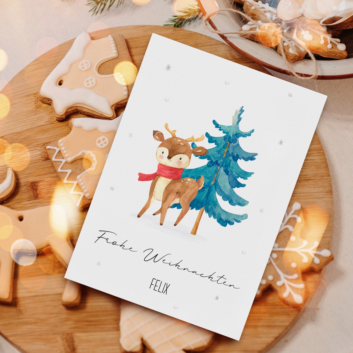 Weihnachtsszene mit Personalisierte Weihnachtskarte Reh & Winterwald A6, zeigt ein Reh mit Tannenbaum, umgeben von Lebkuchen, auf Holzbrett.