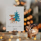 Personalisierte Weihnachtskarte Reh & Winterwald A6 zeigt ein Reh mit rotem Schal vor einem Weihnachtsbaum, begleitet von einem glänzenden Hirschkerzenhalter auf rustikalem Tisch.