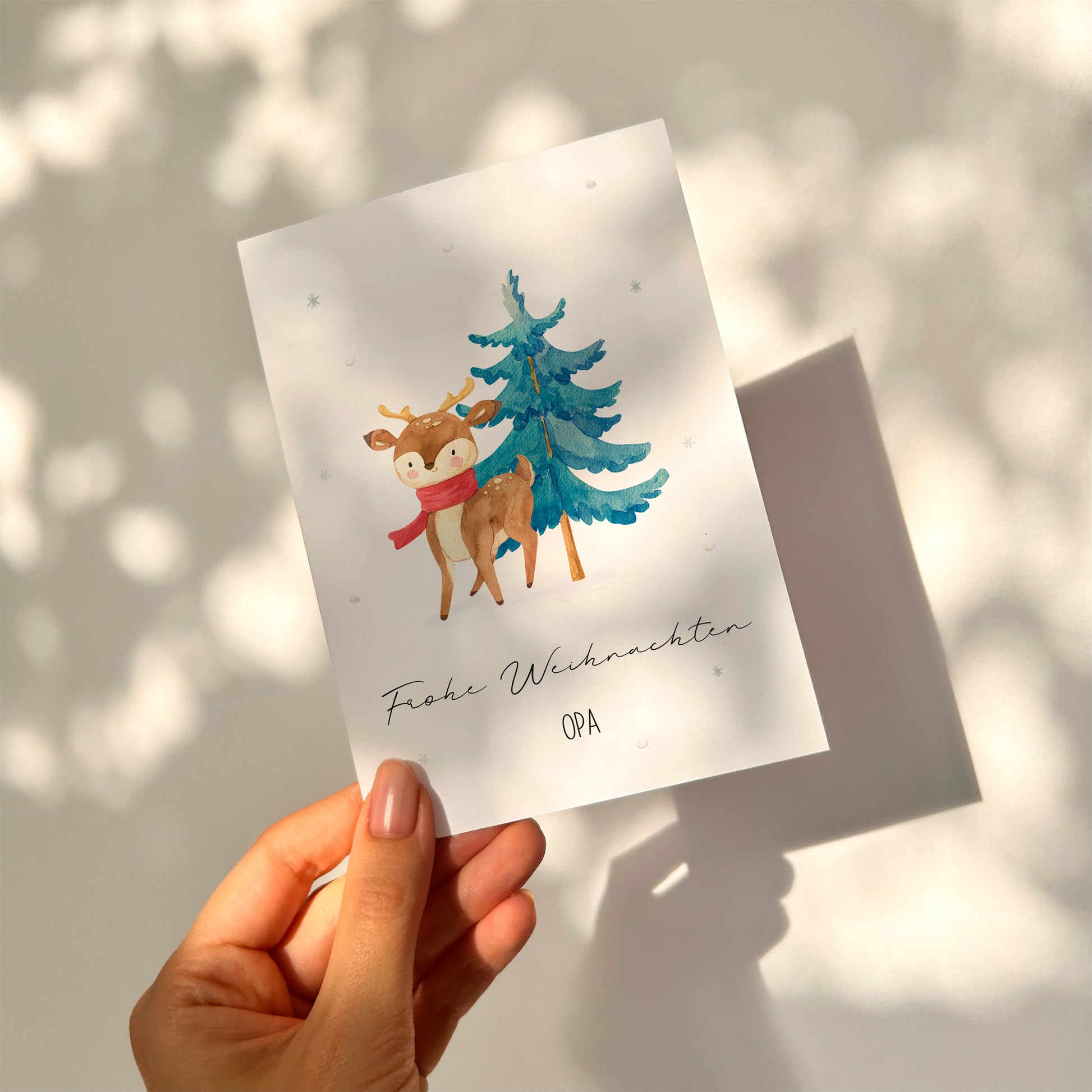 Eine Hand hält die Personalisierte Weihnachtskarte Reh & Winterwald A6 mit einem Reh in rotem Schal vor einem Baum und Frohe Weihnachten Opa in Schrift.