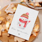 Personalisierte Weihnachtskarte Schneemann A6 mit handgemaltem Schneemann, rotem Schal, winterlicher Mütze. Umgeben von Lebkuchenplätzchen und Lichterdekoration auf Holzbrett.