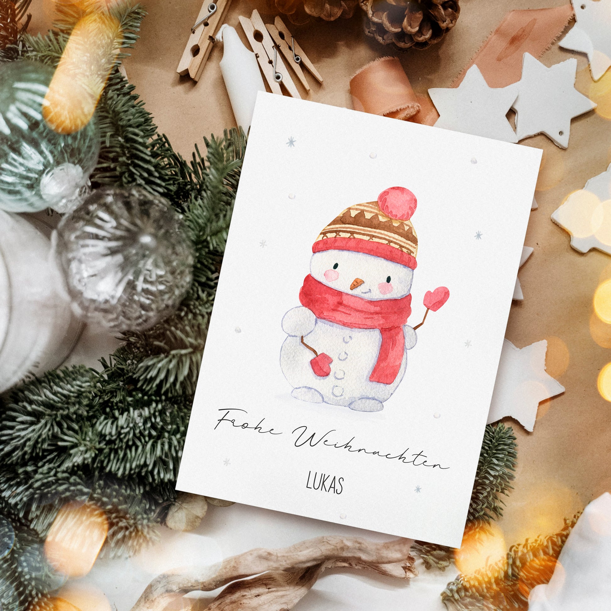 Personalisierte Weihnachtskarte Schneemann A6 mit Aquarell-Schneemann, Frohe Weihnachten und Name Lukas. Umgeben von festlichen Dekorationen wie Tannenzweigen und Holzsternen.