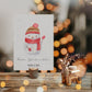 Personalisierte Weihnachtskarte Schneemann A6 auf rustikalem Holz, mit handgemaltem Schneemannmotiv, daneben goldener Rentierkerzenhalter und großer Tannenzapfen, bokeh Lichter im Hintergrund.
