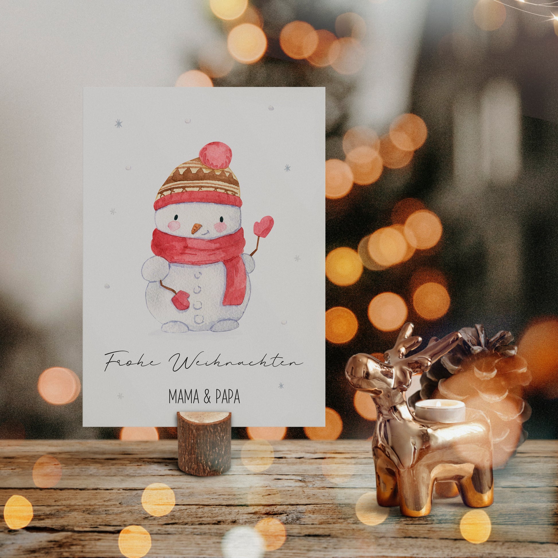 Personalisierte Weihnachtskarte Schneemann A6 auf rustikalem Holz, mit handgemaltem Schneemannmotiv, daneben goldener Rentierkerzenhalter und großer Tannenzapfen, bokeh Lichter im Hintergrund.