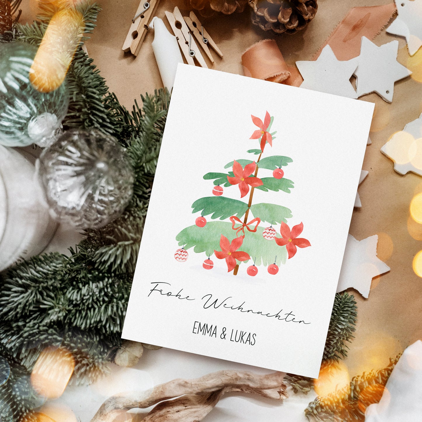 Personalisierte Weihnachtskarte Weihnachtsbaum A6 mit Aquarell-Tannenzweig, roten Blüten und Baubles, umgeben von natürlichen und handgemachten Dekorationen. Text: Frohe Weihnachten und EMMA & LUKAS.