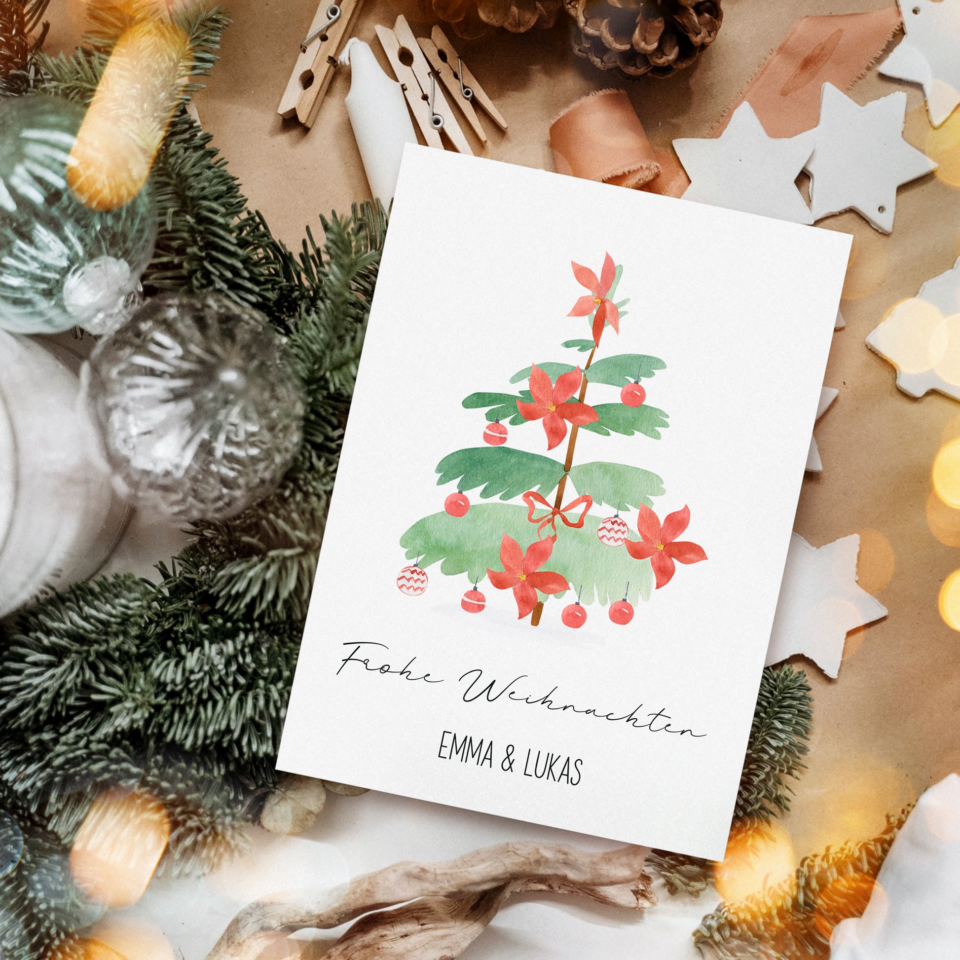 Personalisierte Weihnachtskarte Weihnachtsbaum A6 mit Aquarell-Tannenzweig, roten Blüten und Baubles, umgeben von natürlichen und handgemachten Dekorationen. Text: Frohe Weihnachten und EMMA & LUKAS.