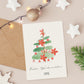 Personalisierte Weihnachtskarte Weihnachtsbaum A6 zeigt handgemalten Weihnachtsbaum mit Poinsettien, individuell anpassbar, inklusive Umschlagoption, umgeben von weihnachtlichen Dekorationen wie Sternen und Lichtern.