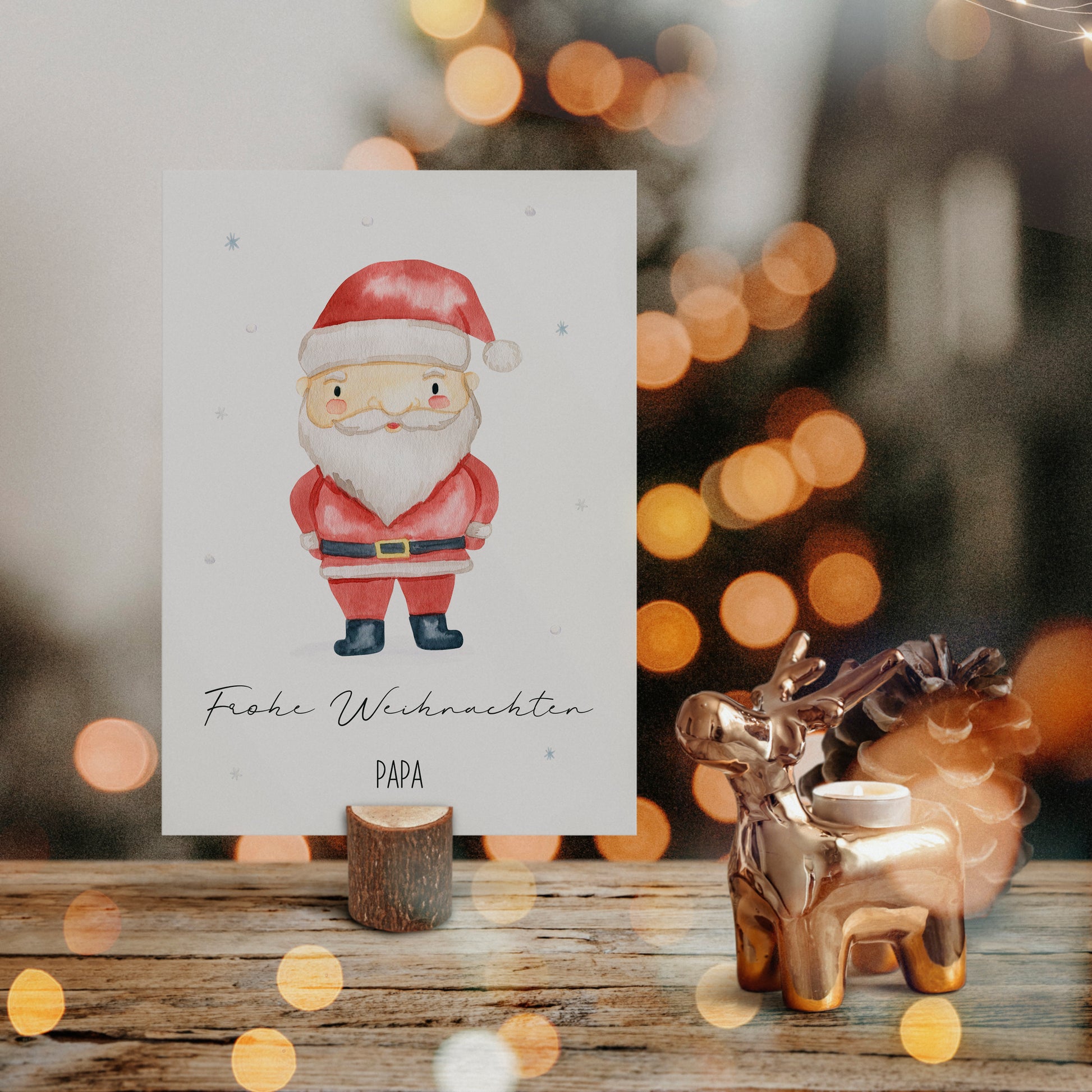 Personalisierte Weihnachtskarte Weihnachtsmann A6 in einem kleinen Holzständer, begleitet von einem goldenen Rentier-Teelichthalter und Tannenzapfen auf rustikalem Holz.