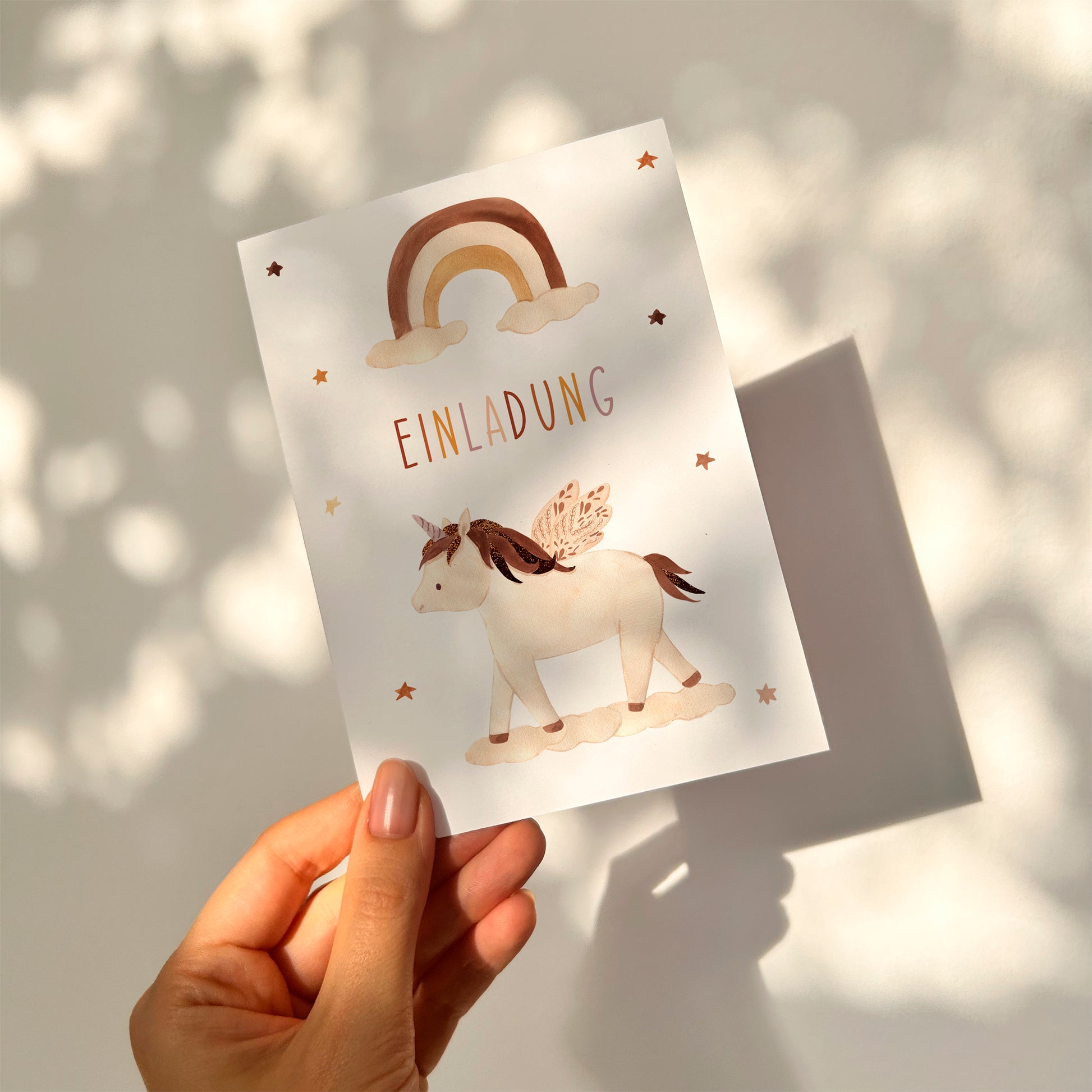 Einladung Kindergeburtstag Einhorn Boho A6: Hand hält eine Einladungskarte mit Regenbogen, Einhorn und Sternenmotiven, geeignet für Kindergeburtstage. Hochwertiges, mattes Naturpapier.