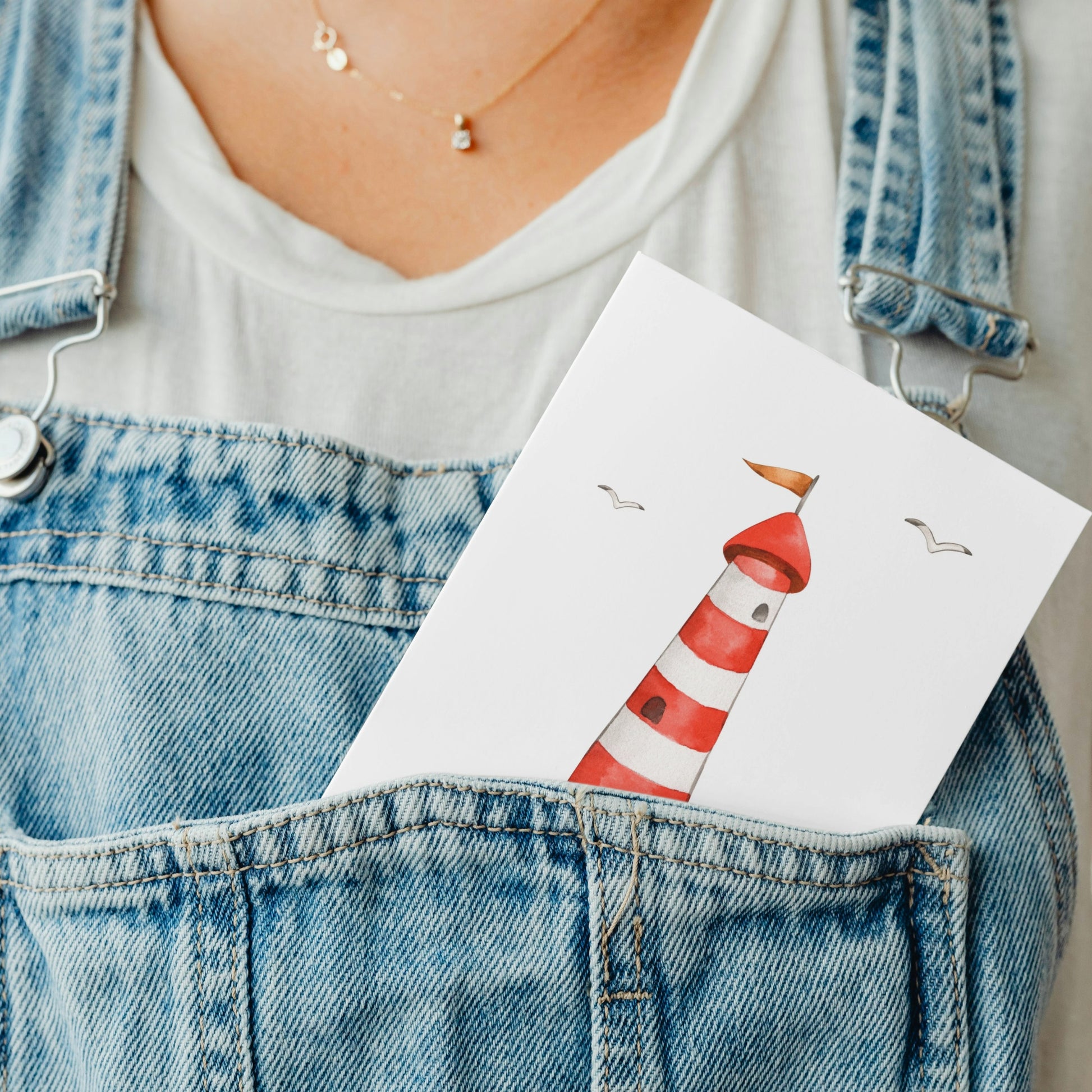 Person trägt blaue Latzhose mit weißem T-Shirt; in der Tasche steckt die Postkarte Leuchtturm A6 mit Leuchtturm-Illustration und Möwen. Goldene Kette sichtbar.