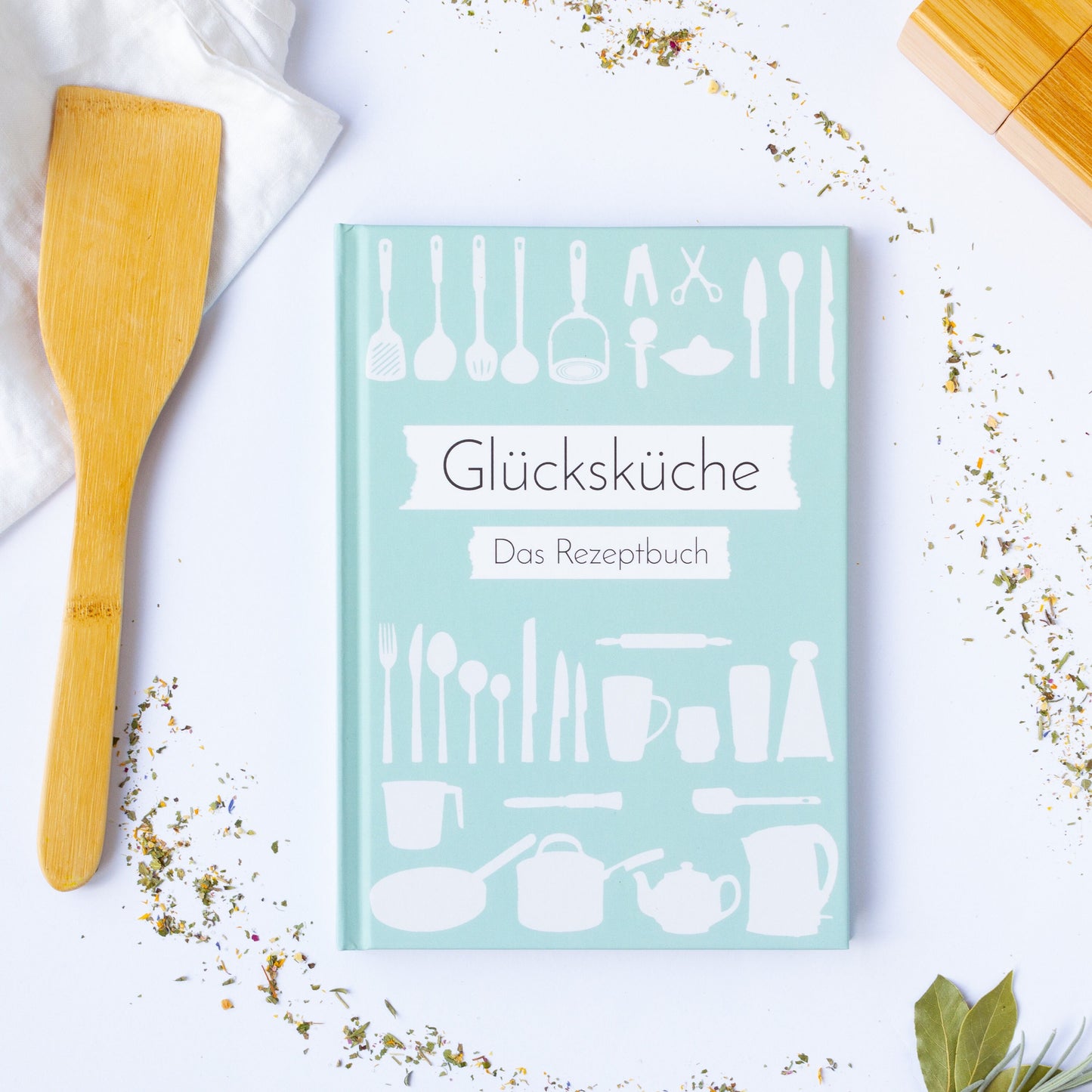 Rezeptbuch zum Selberschreiben A5 Hardcover Glücksküche, grün-weißes Cover, ideal für kreative Rezeptideen. Kräuter und Holzlöffel daneben.