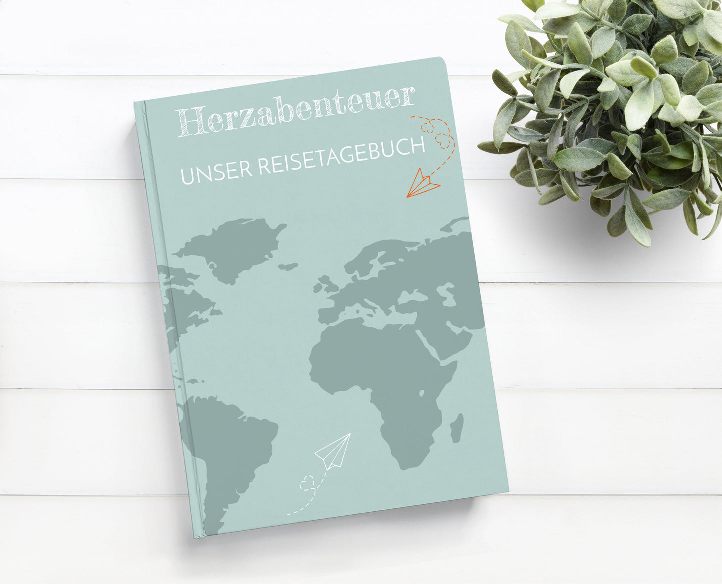 Reisetagebuch für Paare Herzabenteuer A5 Hardcover auf einem Tisch, mit Weltkarte und Papierfliegern auf dem Cover. Text: Unser Reisetagebuch.