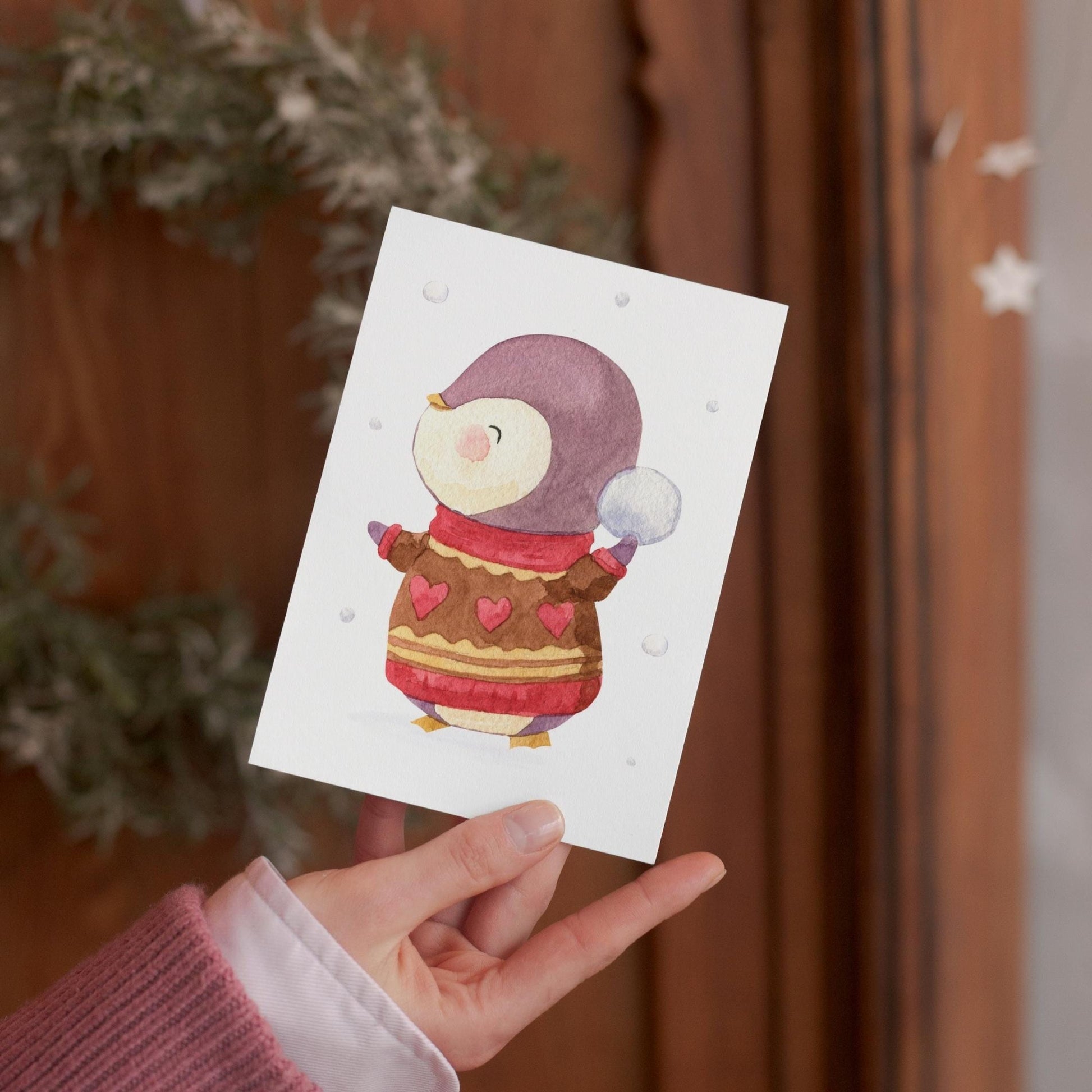 Eine Person hält eine Weihnachtskarte mit einer Aquarell-Illustration von Pinguin, der einen Schneeball wirft.