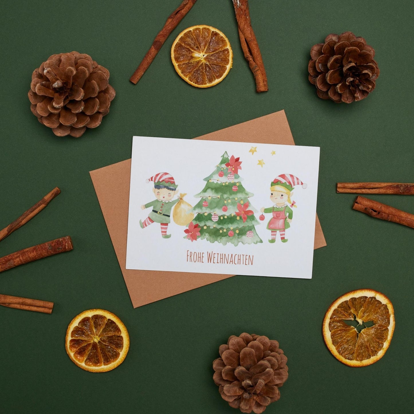 Weihnachtskarte Elfen & Weihnachtsbaum Frohe Weihnachten A6, umgeben von Tannenzapfen und getrockneten Orangenscheiben, mit süßer weihnachtlicher Illustration und Frohe Weihnachten Schriftzug.