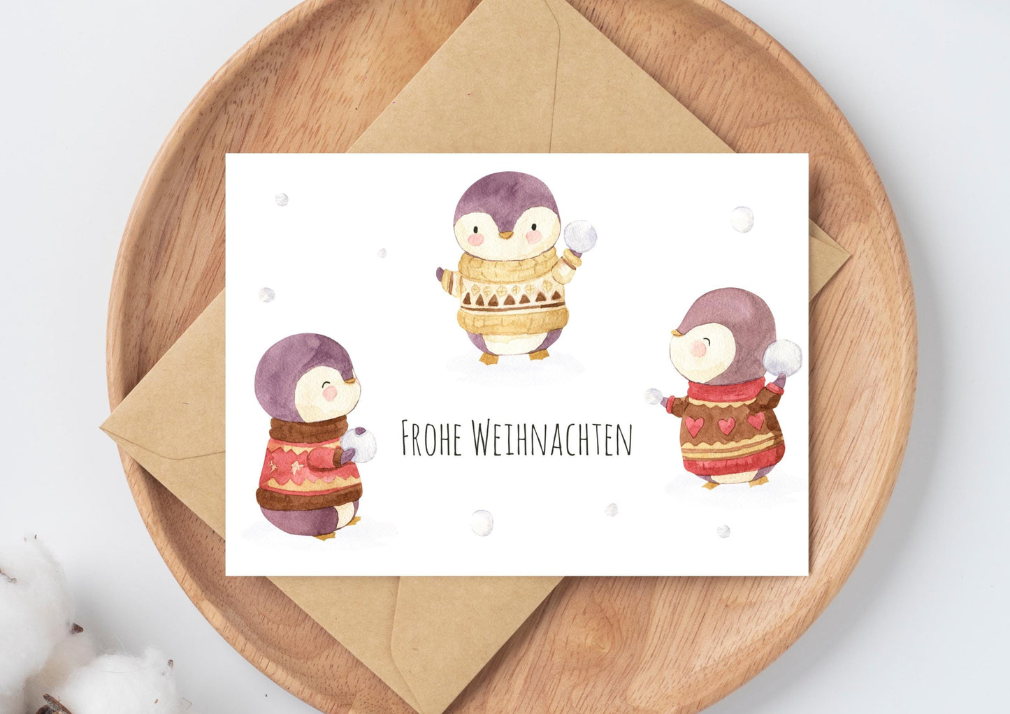 Weihnachtskarte Schneeballschlacht Frohe Weihnachten A6 mit süßer Illustration: Pinguine in Pullovern. Beiliegend ein passender Umschlag auf einem Holztablett.