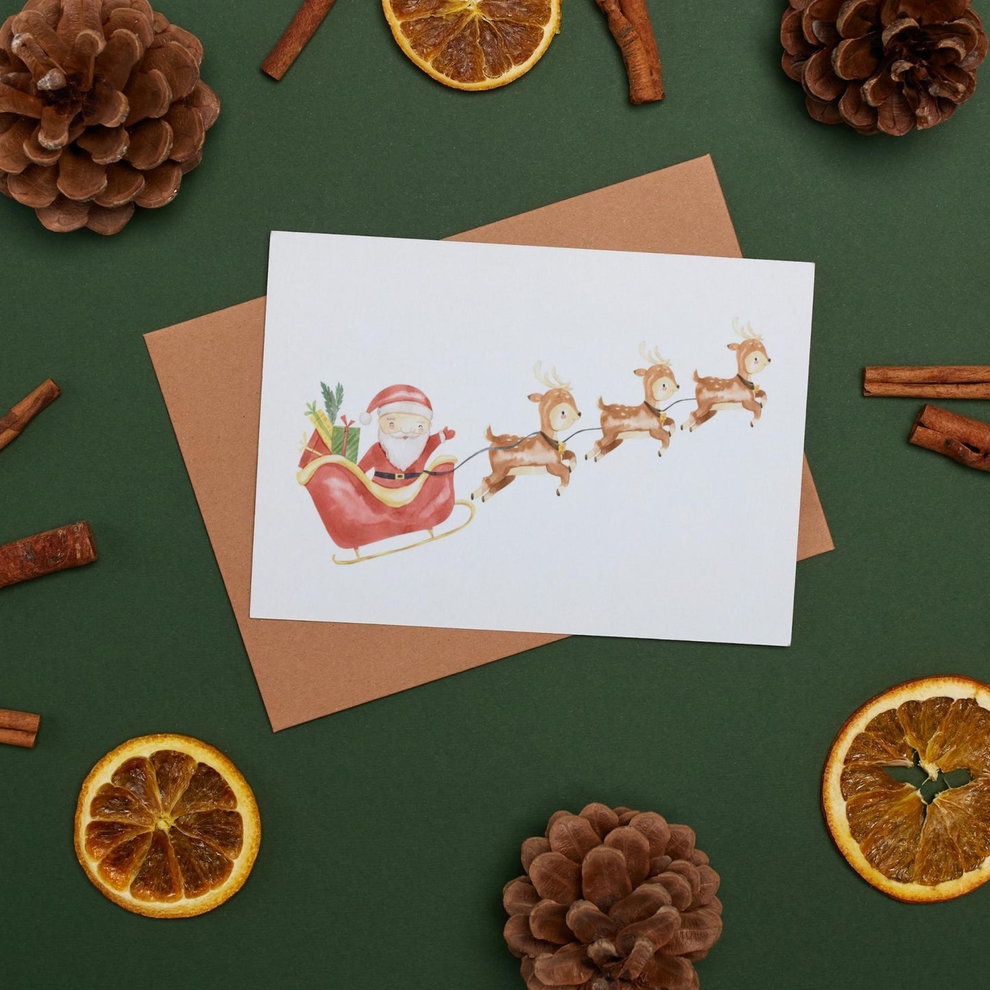 Weihnachtskarte Weihnachtsschlitten A6: Santa Claus und Rentiere. Umgeben von Tannenzapfen und getrockneten Orangenscheiben, auf hochwertigem Naturpapier gedruckt.