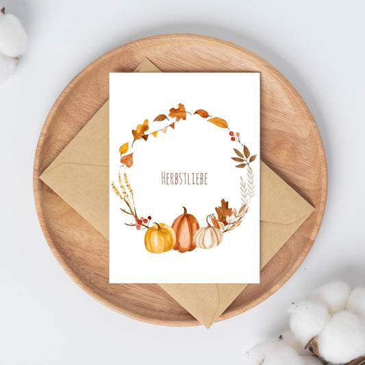 Postkarte Herbstliebe A6 mit herbstlichem Kranz und Kürbis-Motiv, aus 300g Naturpapier, auf einem Teller präsentiert.