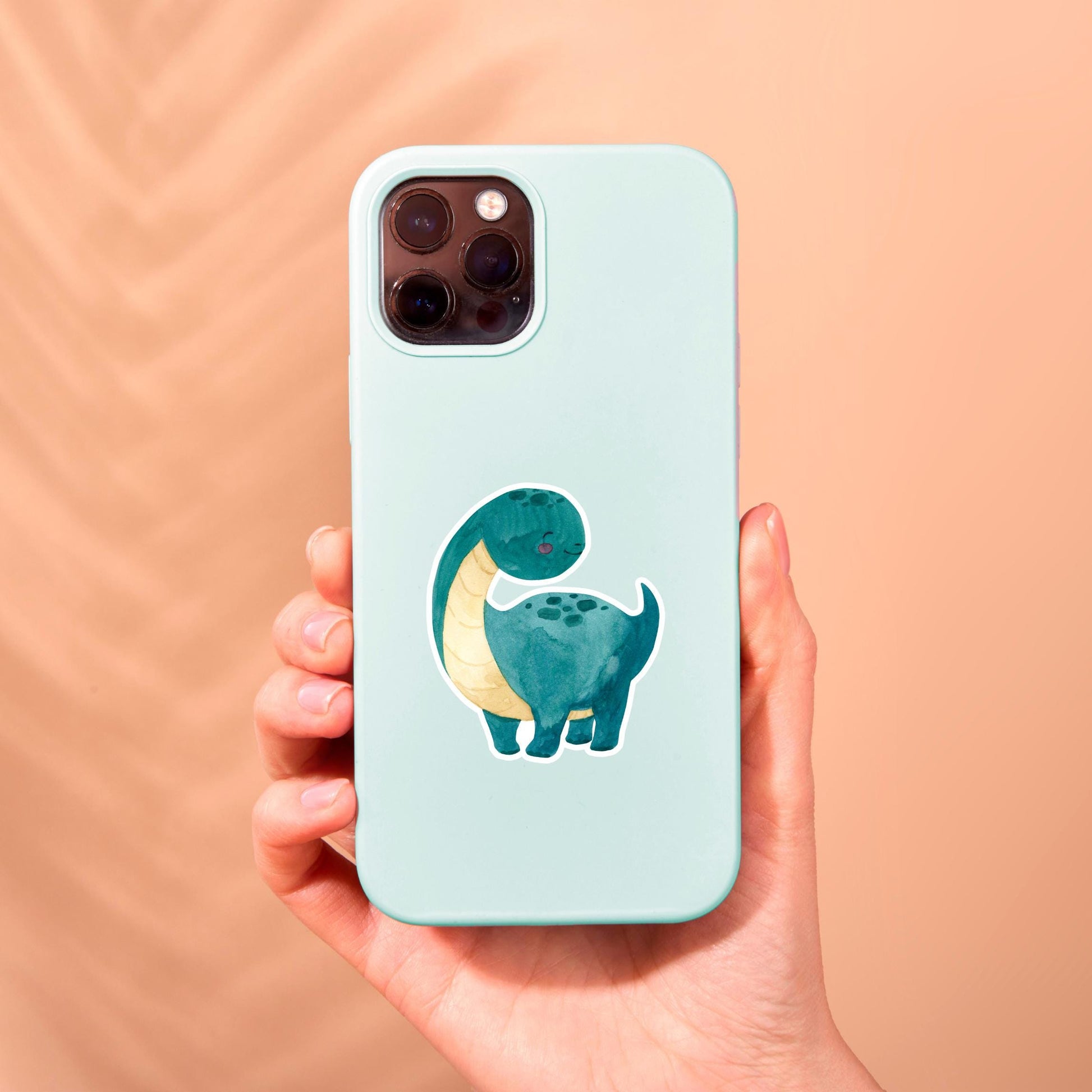 Hand hält Handyhülle mit Sticker Dinosaurier dunkelgrün, handgemalter Cartoon-Dino, Vinyl, spritzfest, auf Handyhülle platziert, ideal für Dino-Fans.