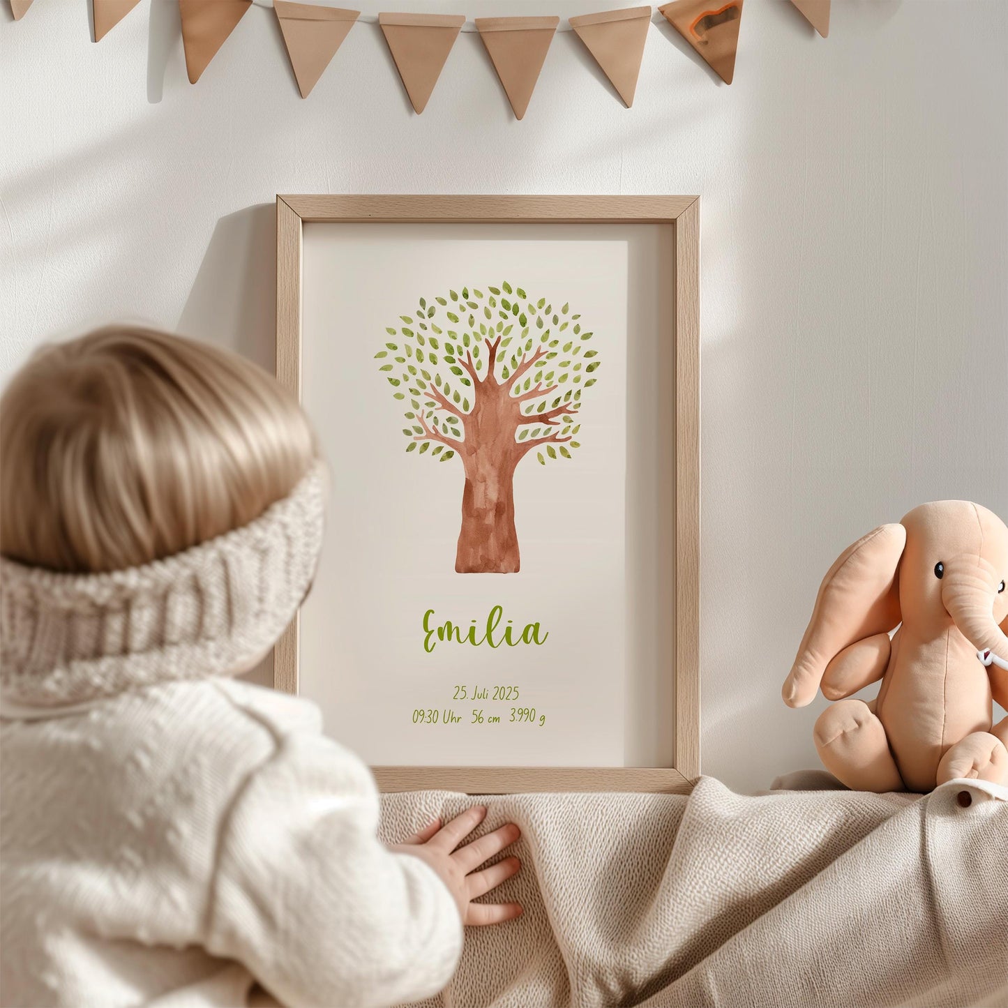 Geburtsposter Baum personalisiert: Kleinkind greift nach gerahmtem Poster mit Baumgrafik und Namen Emilia, daneben ein Elefanten-Plüschtier und eine Wimpelkette.