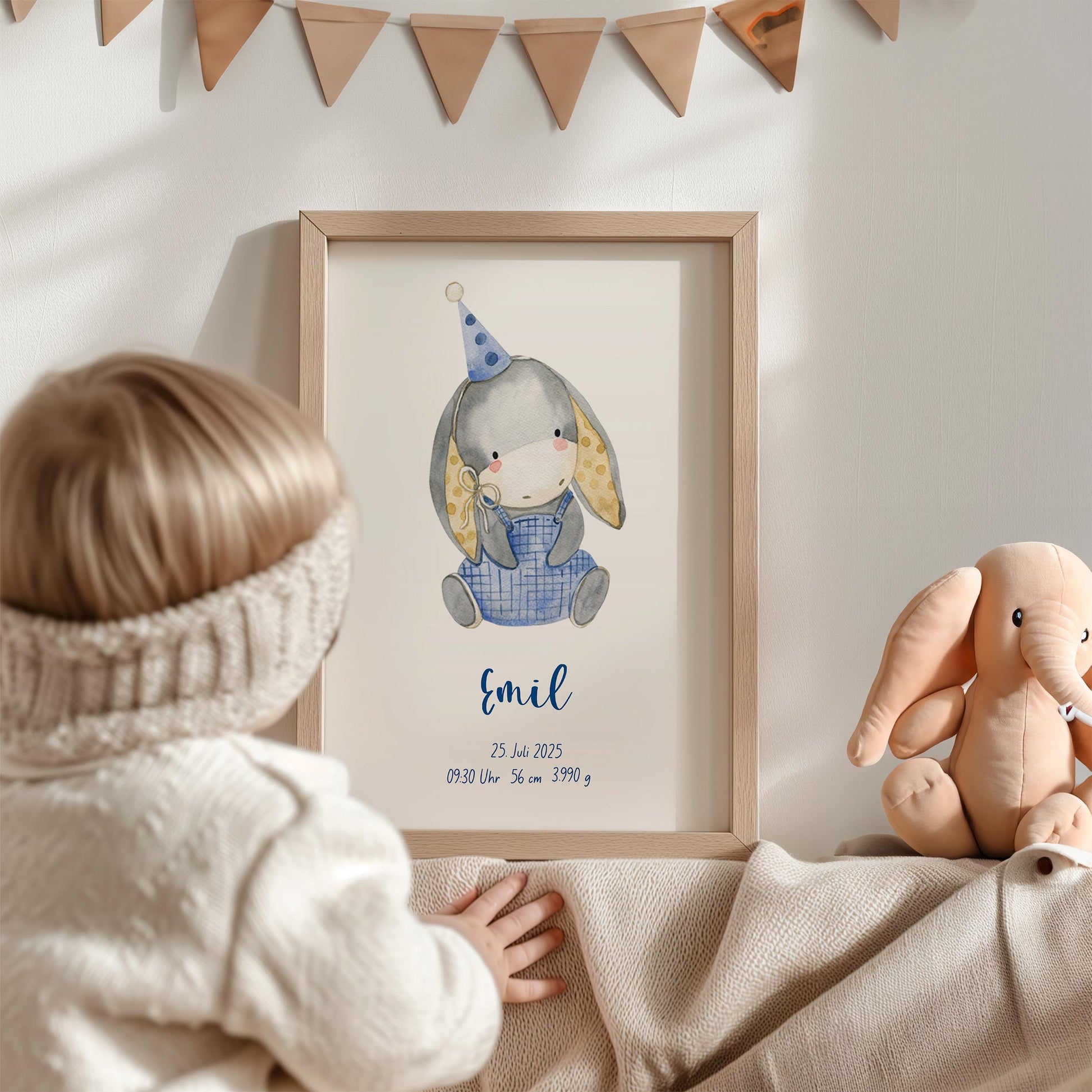 Geburtsposter Esel personalisiert im Holzrahmen zeigt einen grauen Aquarell-Hasen mit Partyhut; daneben ein Elefanten-Plüschtier, Kind betrachtet neugierig das Bild.