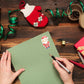 Hände schreiben auf grünem Papier mit einem Sticker Weihnachtsmann. Umgeben von festlichen Dekorationen, ideal für kreative Weihnachtskarten und Geschenke.