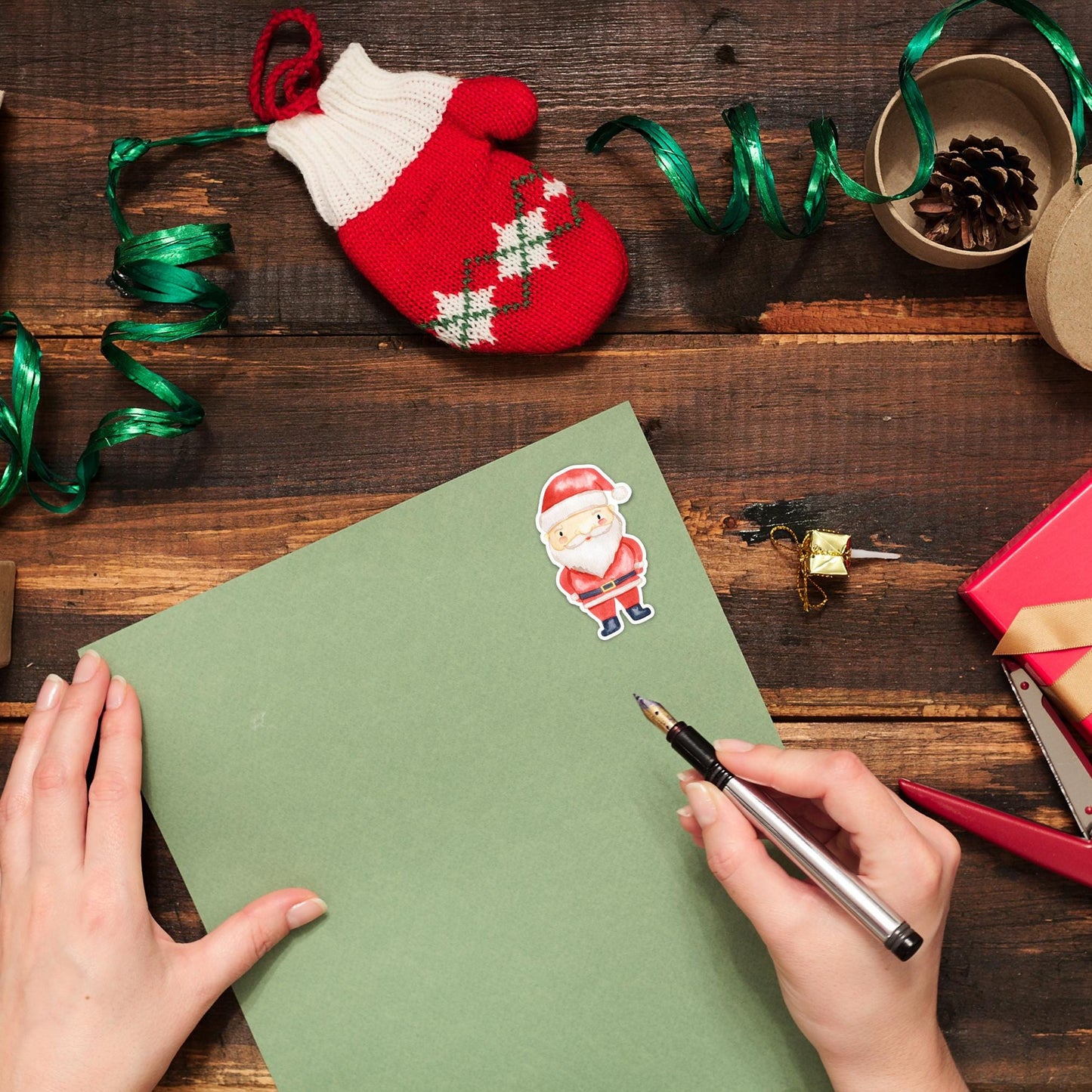 Hände schreiben auf grünem Papier mit einem Sticker Weihnachtsmann. Umgeben von festlichen Dekorationen, ideal für kreative Weihnachtskarten und Geschenke.
