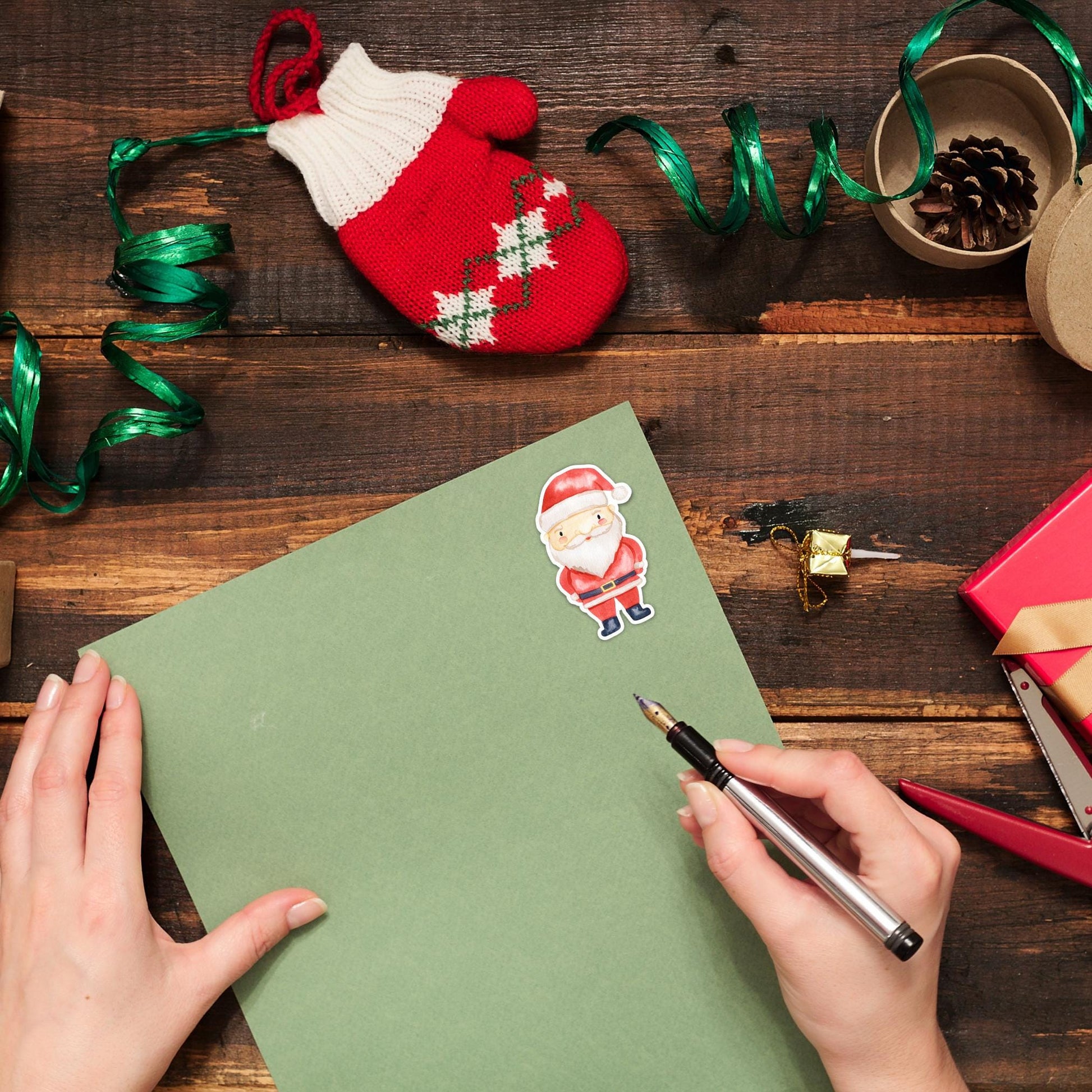 Hände schreiben auf grünem Papier mit einem Sticker Weihnachtsmann. Umgeben von festlichen Dekorationen, ideal für kreative Weihnachtskarten und Geschenke.