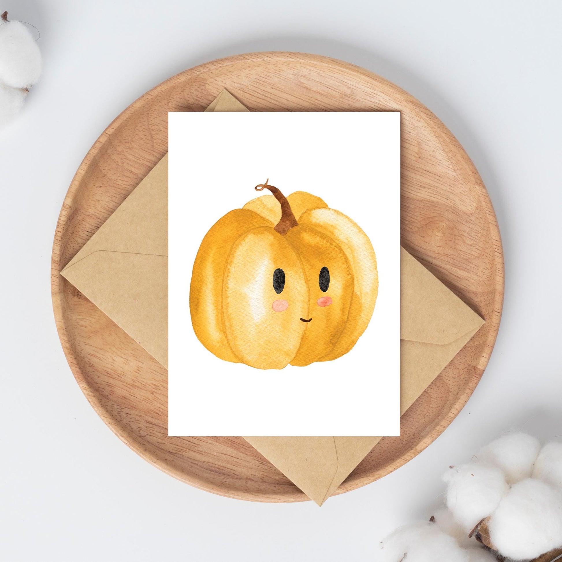 Postkarten Set Kürbis A6: Runde Holzschale mit Kraftpapierumschlag und Karte, zeigt goldgelben Kürbis mit Gesicht, umgeben von Baumwollblüten auf weißem Tisch.