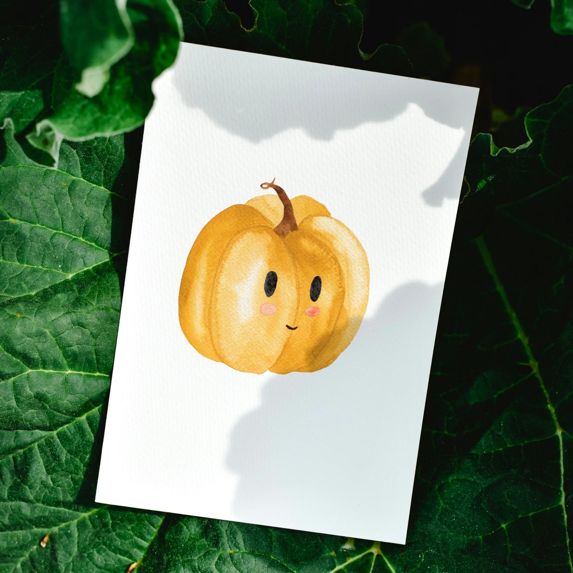 Postkarten Set Kürbis A6 zeigt eine Karte mit einem lächelnden Kürbis auf weißem Papier, platziert auf dunklen Blättern. Ideal für Herbstgrüße.