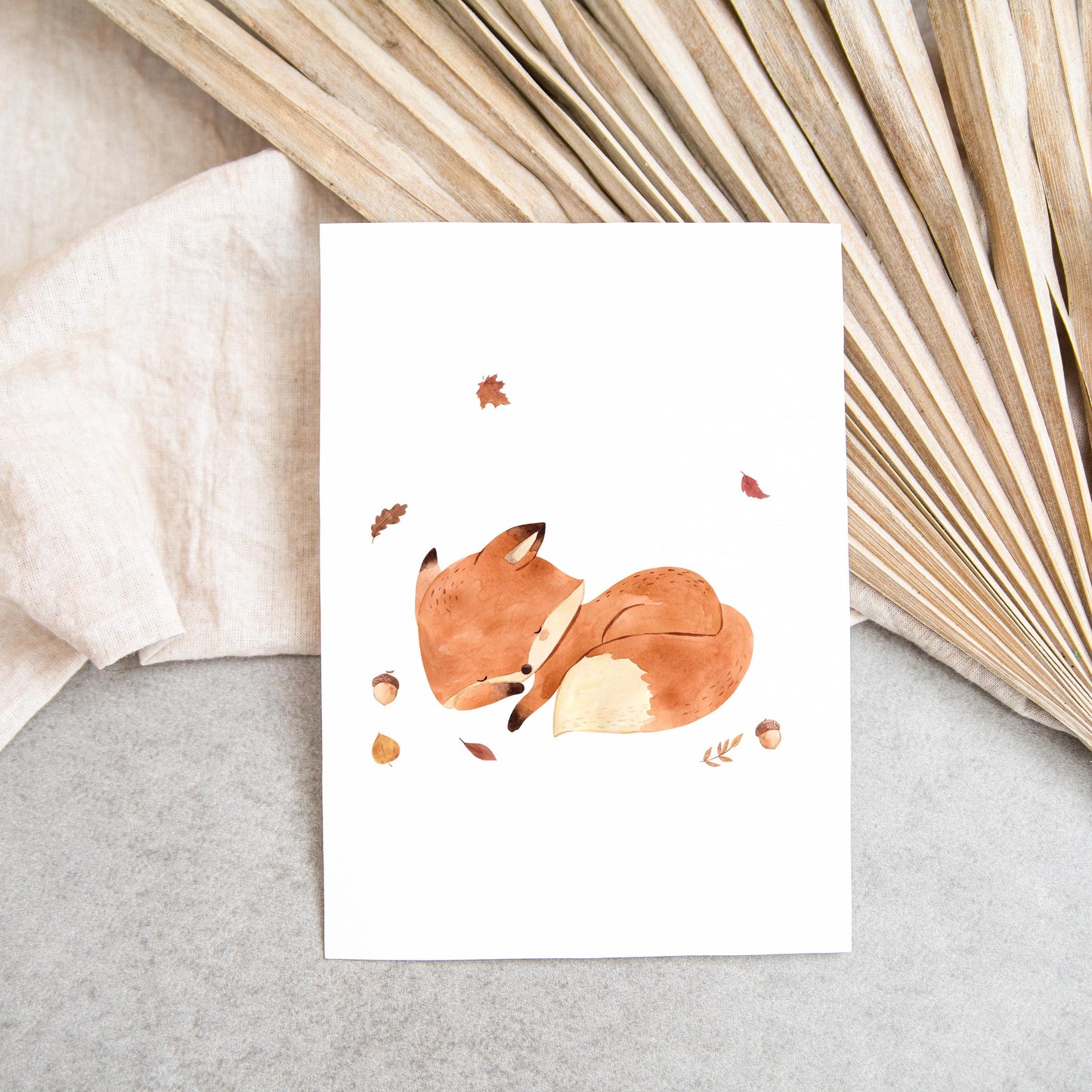 Karte Fuchs & Herbstlaub A6 mit einem schlafenden Fuchs in Aquarell auf weißem Papier, umgeben von Blättern und Eicheln, auf grauem Untergrund.