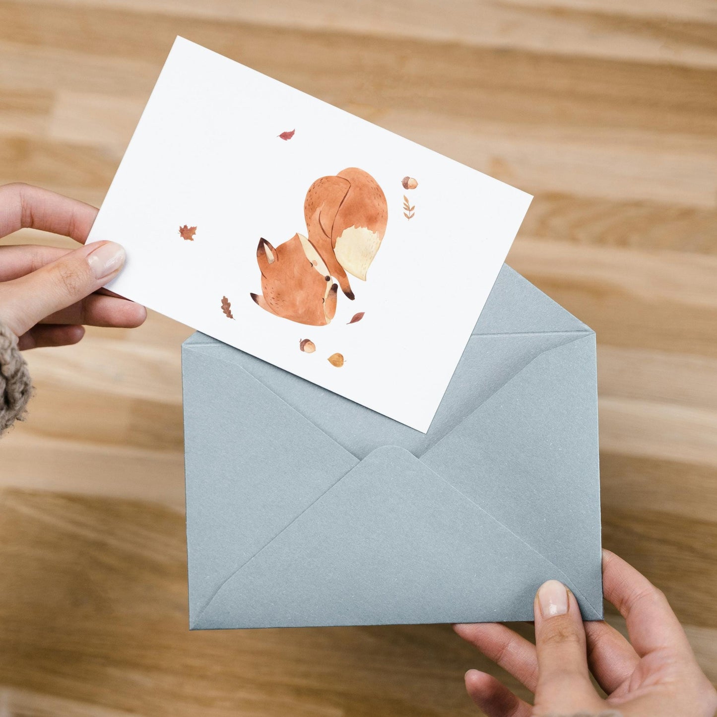 Karte Fuchs & Herbstlaub A6: Eine Person zieht eine Postkarte mit schlafendem Fuchs, Herbstlaub und Eicheln aus einem blauen Umschlag auf Holzoberfläche heraus.
