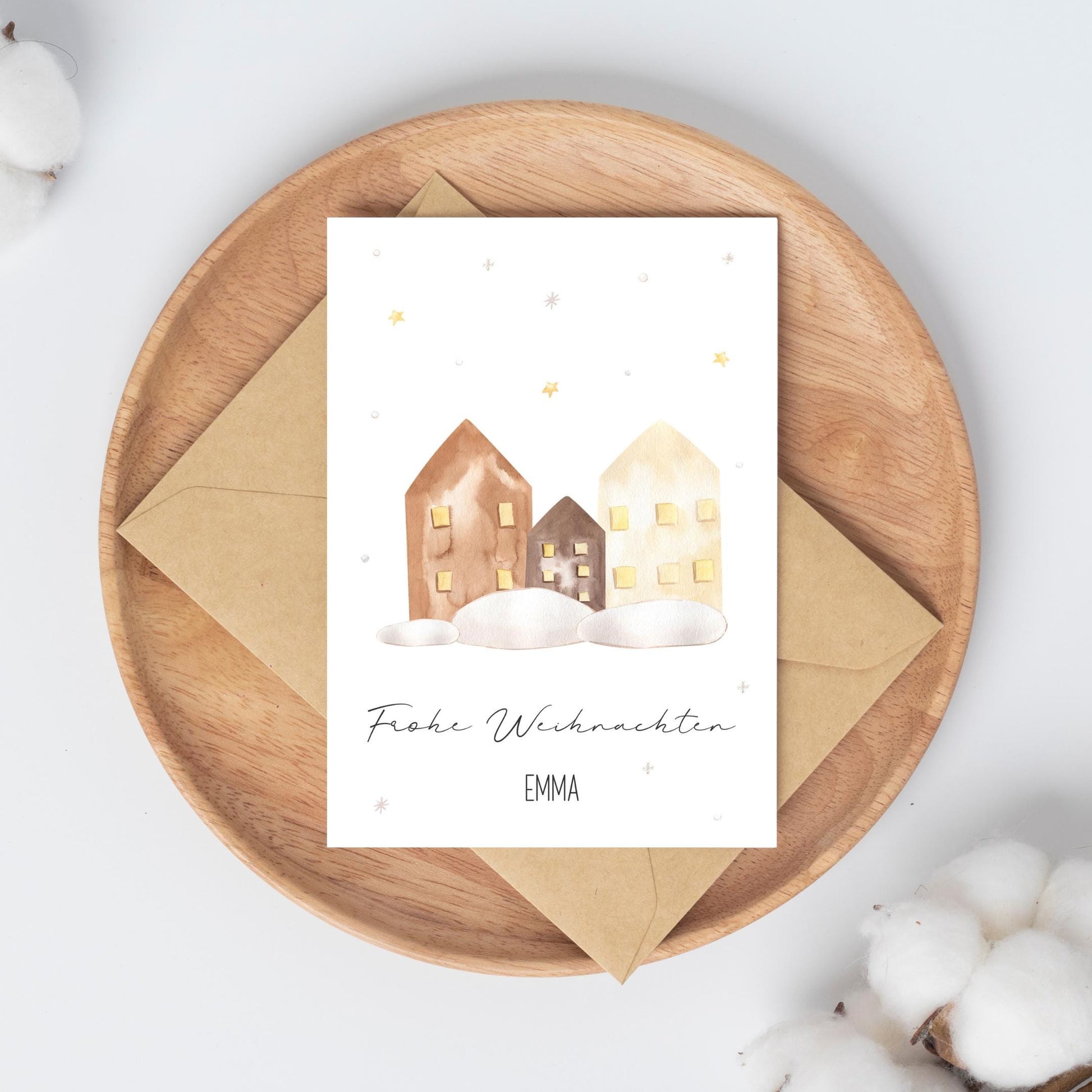 Personalisierte Weihnachtskarte Häuser A6 mit Aquarellillustration von drei verschneiten Häusern und goldenen Sternen. Frohe Weihnachten und Name EMMA. Auf Holztablett mit Umschlag.