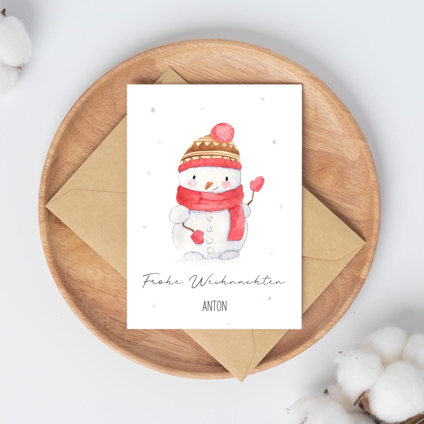 Personalisierte Weihnachtskarte Schneemann A6, zeigt handgemalten Schneemann mit Herz-Stöcken und Frohe Weihnachten Anton. Liegt auf rundem Holztablett mit Umschlag darunter.