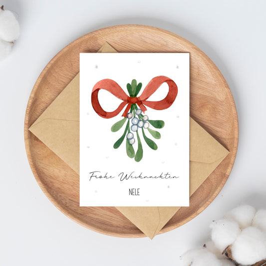 Personalisierte Weihnachtskarte Mistelzweig A6: Runde Holzplatte mit Karte, Mistelzweig-Aquarell, roter Schleife, Frohe Weihnachten Nele Text, daneben ein Umschlag.