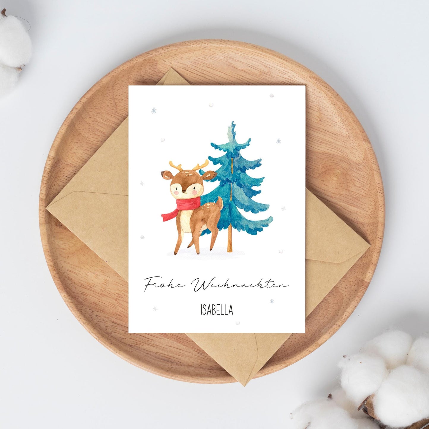 Personalisierte Weihnachtskarte Reh & Winterwald A6 auf Holztablett, zeigt Reh mit Schal und Baum, daneben Umschlag und Baumwollzweige.