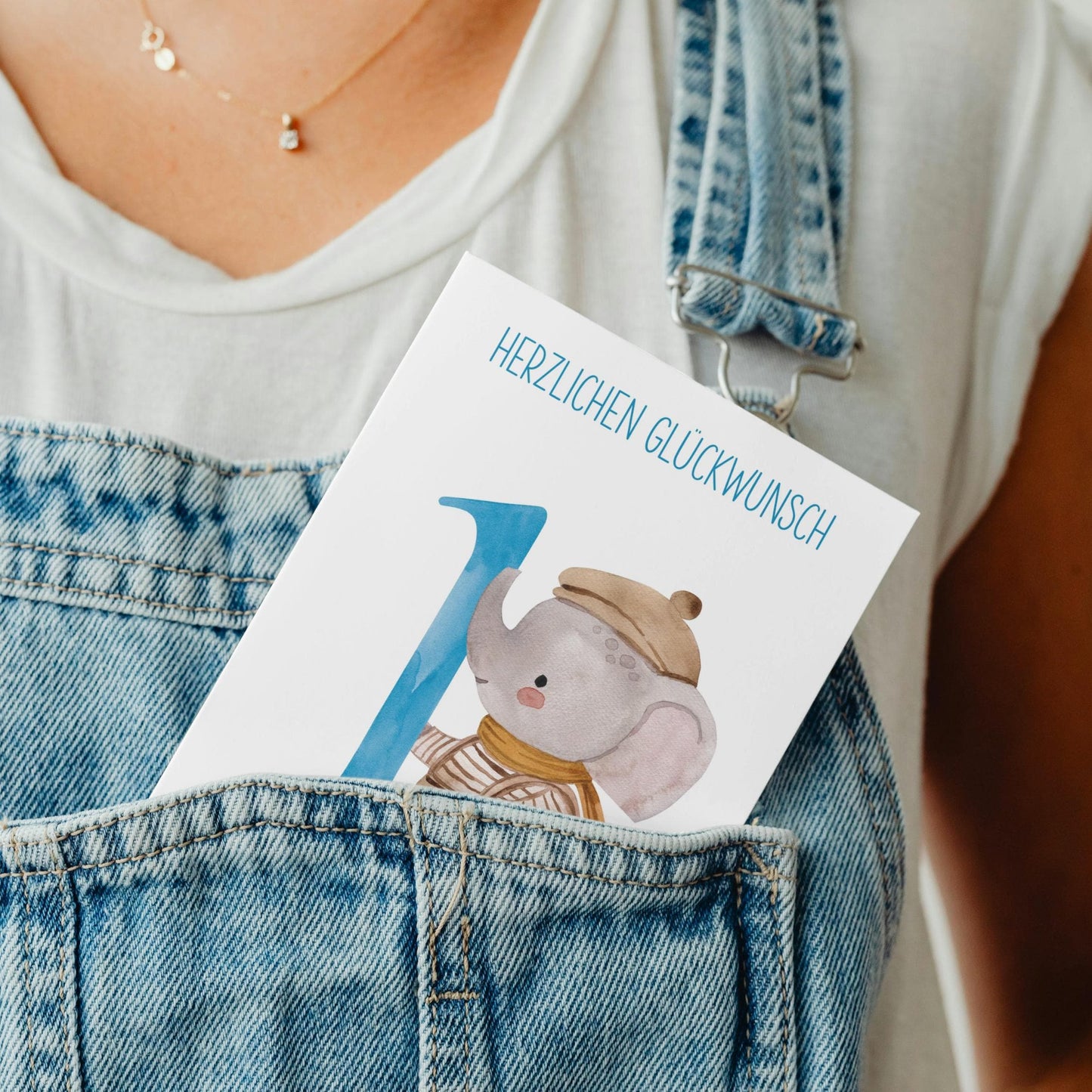 Person trägt blaue Latzhose mit Personalisierte Geburtstagskarte Elefant A6 im Frontfach, zeigt Herzlichen Glückwunsch, Nummer 1, Elefant Illustration.