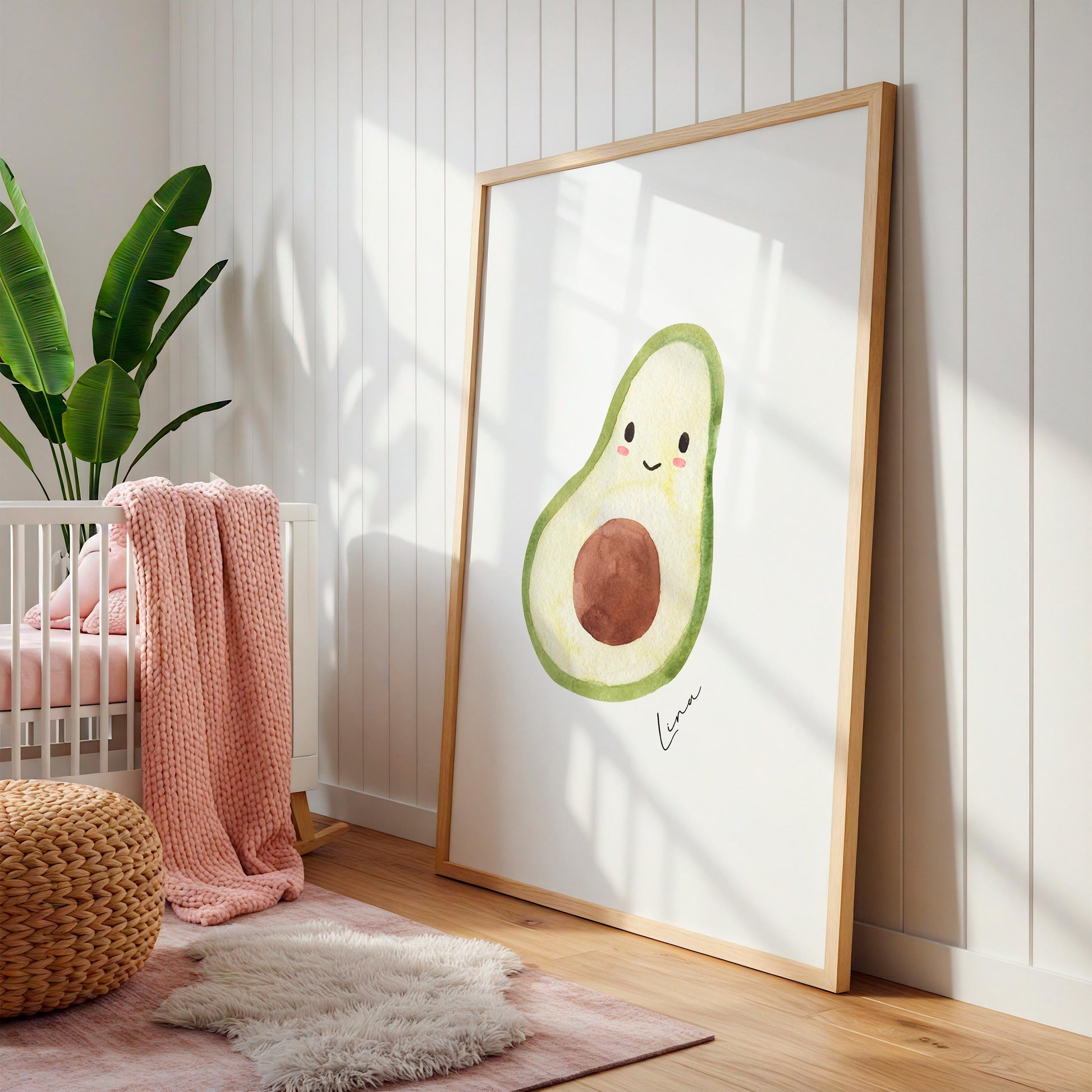 Namensposter Avocado A3 & A4: Ein großes Holzrahmenbild mit fröhlicher Avocado-Illustration und „Lina“, umgeben von Babyzimmerdekorationen wie einem weißen Kinderbett und Pflanzen.