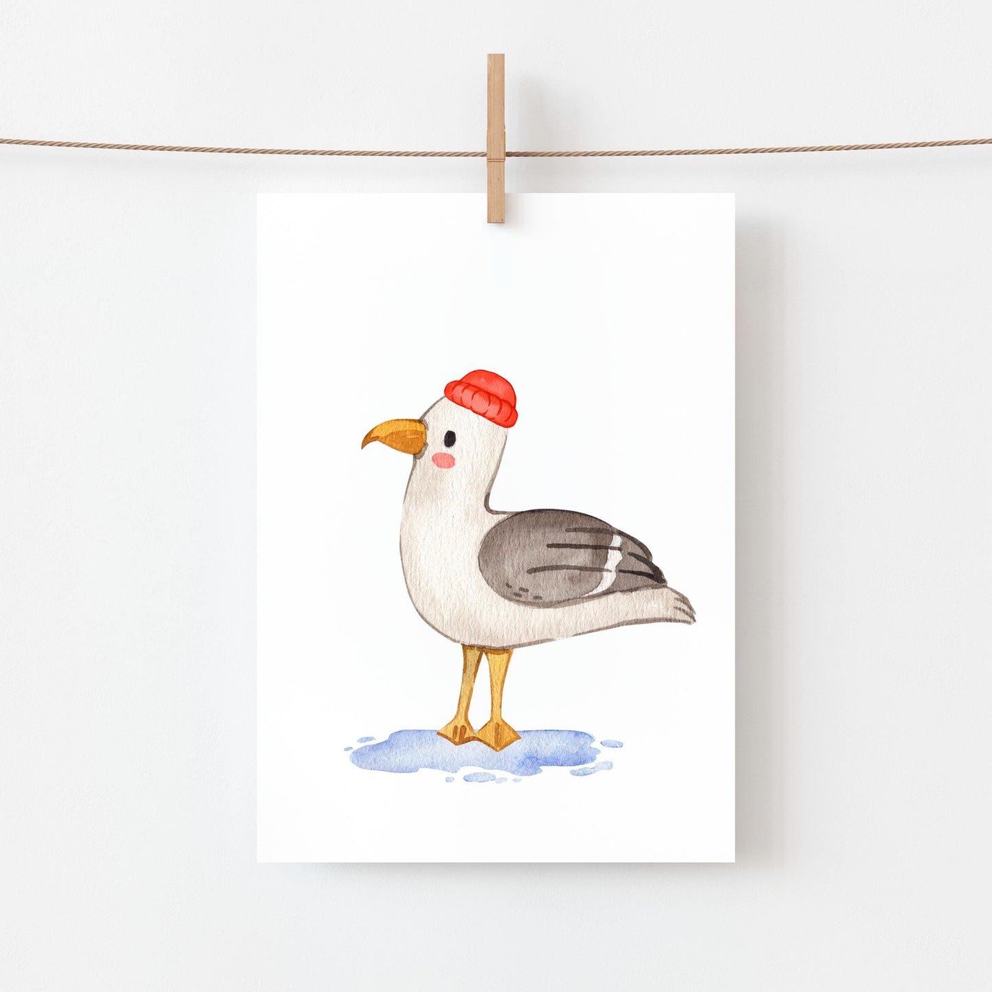 Postkarte Möwe A6 zeigt eine Möwe im Aquarellstil auf Naturpapier, mit gelbem Schnabel, grauen Flügeln und roter Mütze, aufgehängt an einer Kordel.