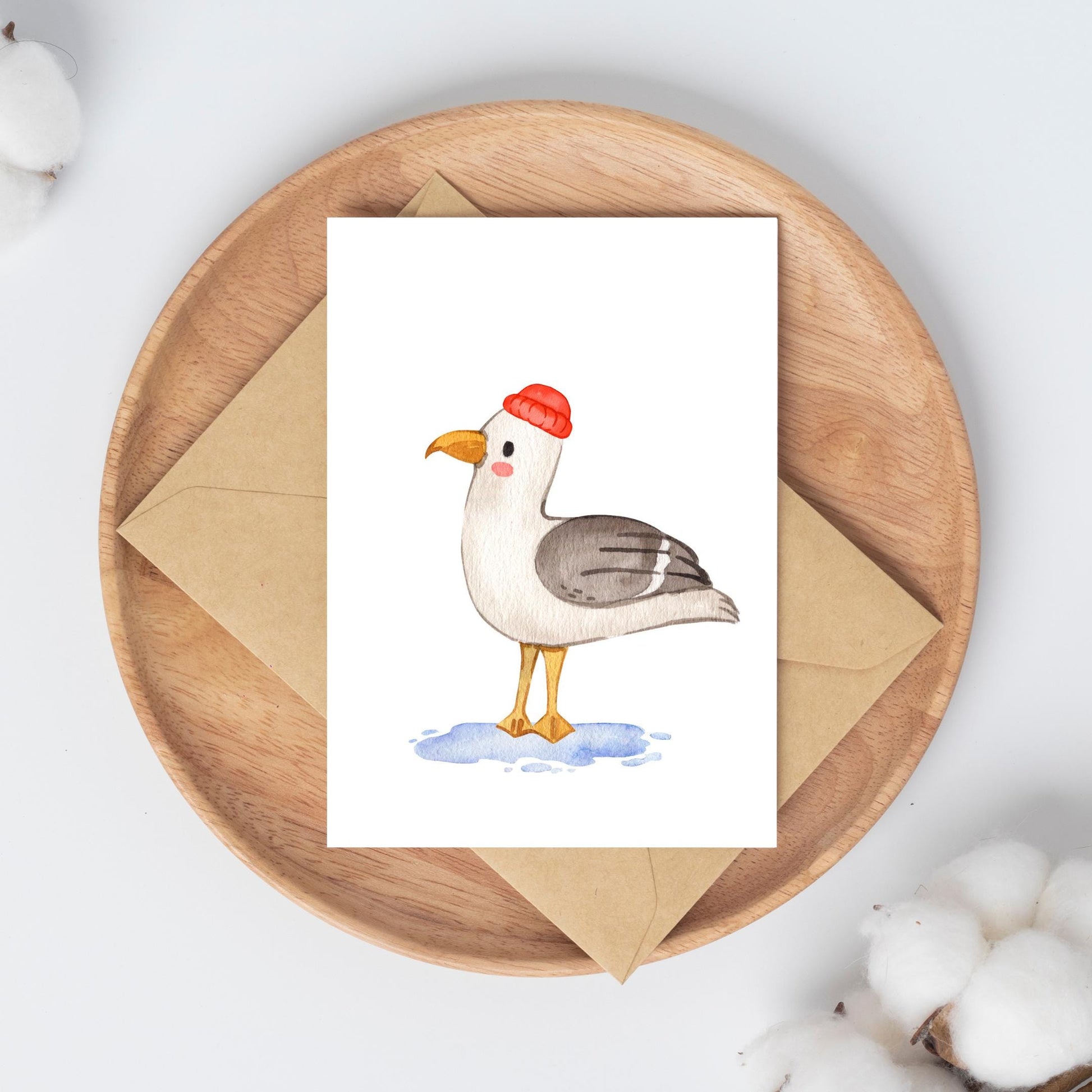 Postkarte Möwe A6 auf einem Holztablett, zeigt eine Möwe mit roter Mütze in Aquarell, ruht auf einem Kraftpapierumschlag, umgeben von Baumwollkapseln.