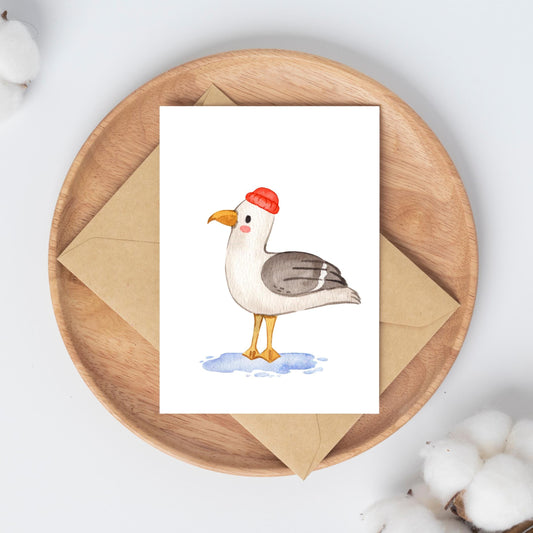 Postkarte Möwe A6 auf einem Holztablett, zeigt eine Möwe mit roter Mütze in Aquarell, ruht auf einem Kraftpapierumschlag, umgeben von Baumwollkapseln.
